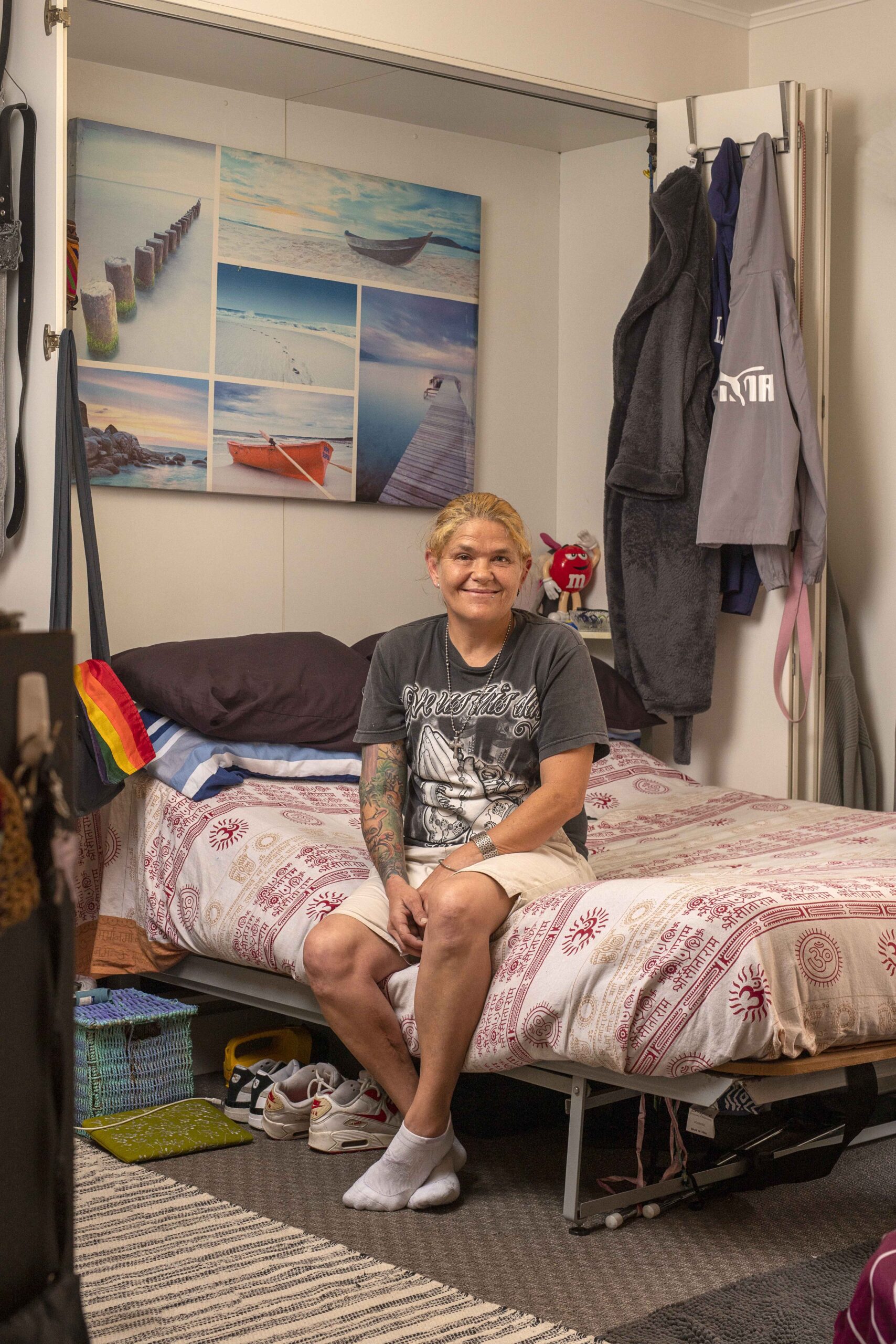 A person with light skin and short, blonde hair sits on a bed with a patterned blanket. They wear a dark graphic t-shirt and shorts, smiling. The room features a wall with landscape artwork and various clothing items hanging nearby.