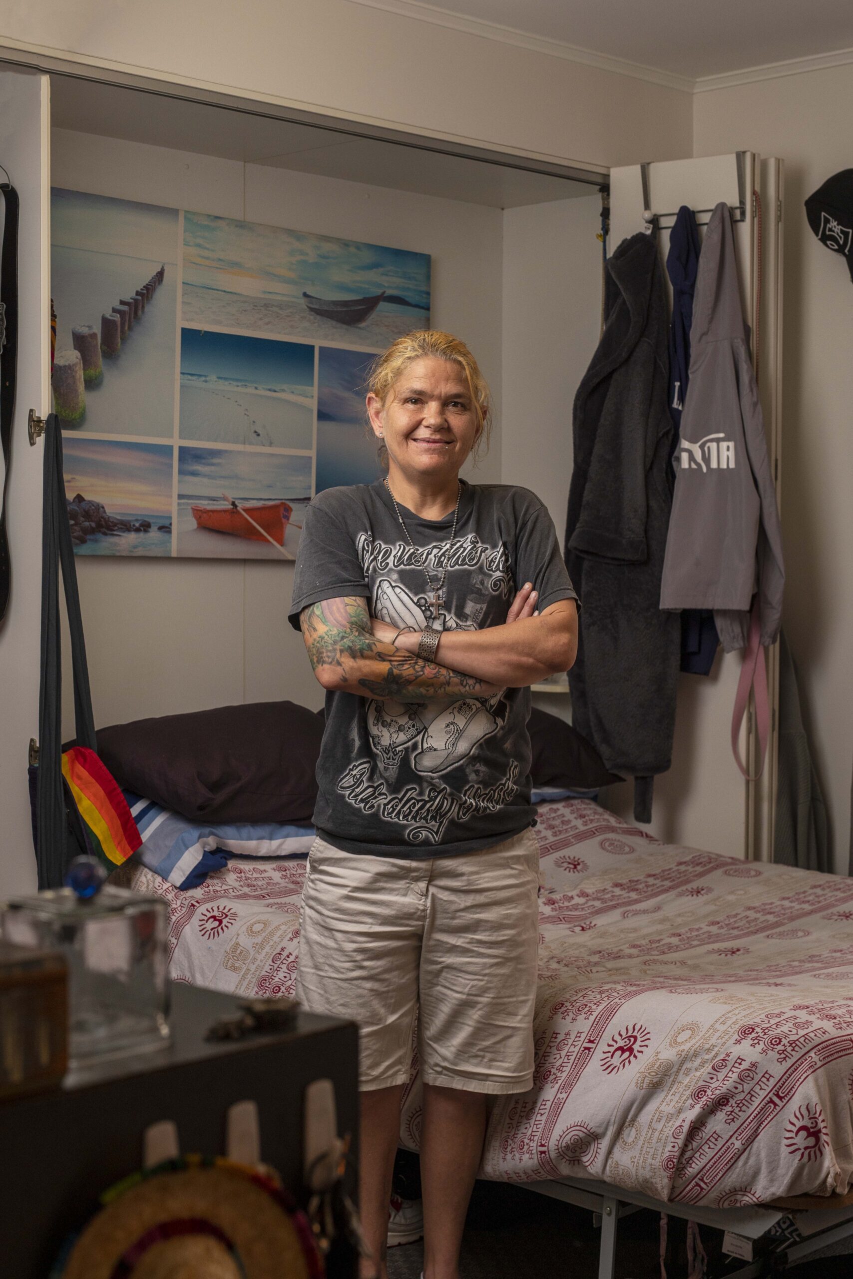 Person with light skin and short blonde hair stands confidently with arms crossed in a bedroom. They wear a dark graphic t-shirt and light-colored pants. The room features a bed with a patterned blanket and various clothing items hanging nearby.