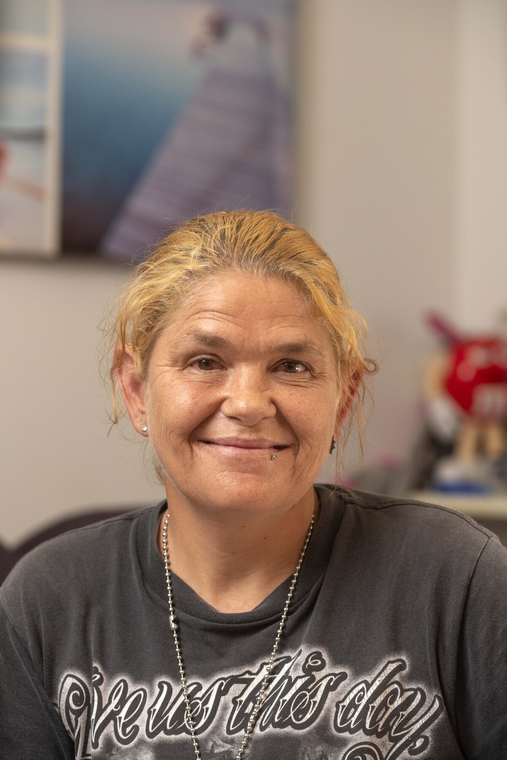 Portrait of a middle-aged woman with light brown skin and blonde hair, wearing a dark gray T-shirt with white lettering. She has a slight smile and a piercing on her lower lip, sitting in an indoor setting with blurred background elements.