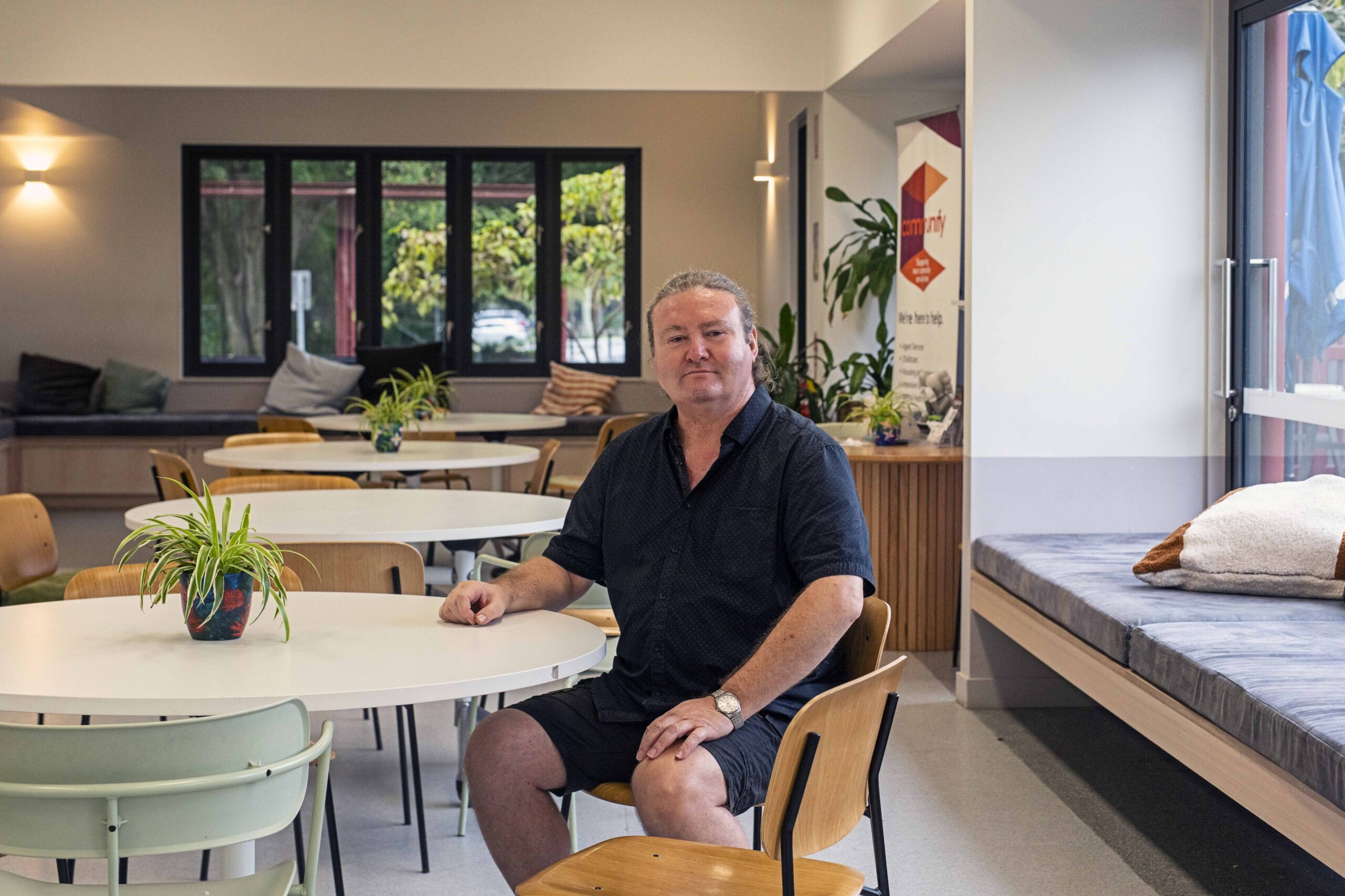 A middle-aged man with light skin is seated at a table in a modern café. He is wearing a black short-sleeve shirt and black shorts. The interior features several tables and plants, with large windows in the background.