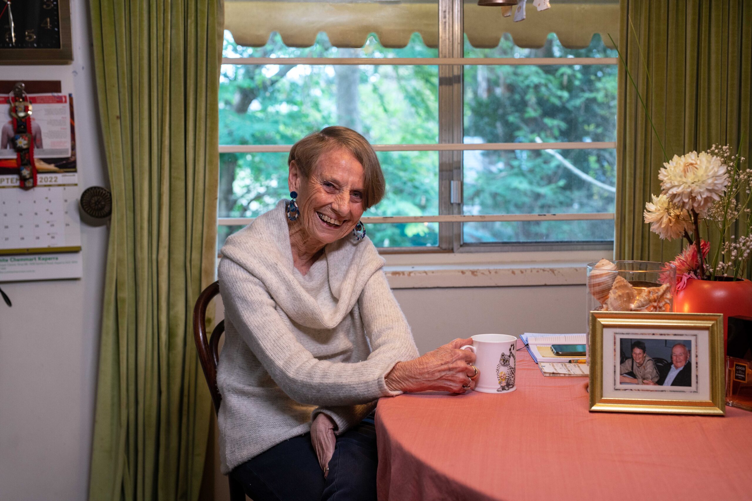 Smiling older woman with short blonde hair wearing a light-colored sweater, seated at a table with a pink tablecloth, holding a mug. In the background, there are green curtains and a window with natural light.