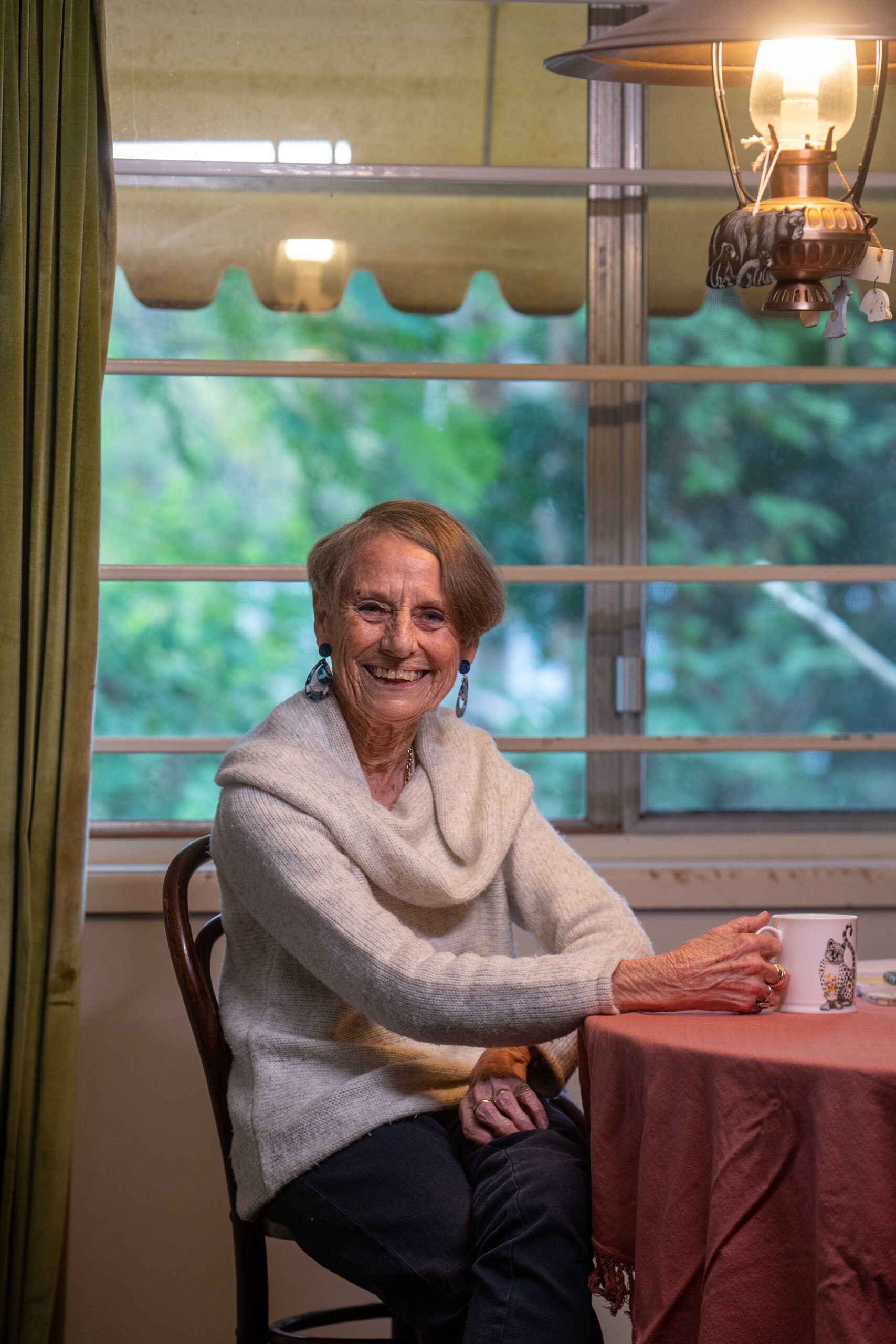 Smiling elderly woman with light skin, wearing a cream-colored sweater, sitting at a table with a pink tablecloth, holding a mug. She is positioned in front of a window with greenery visible outside.