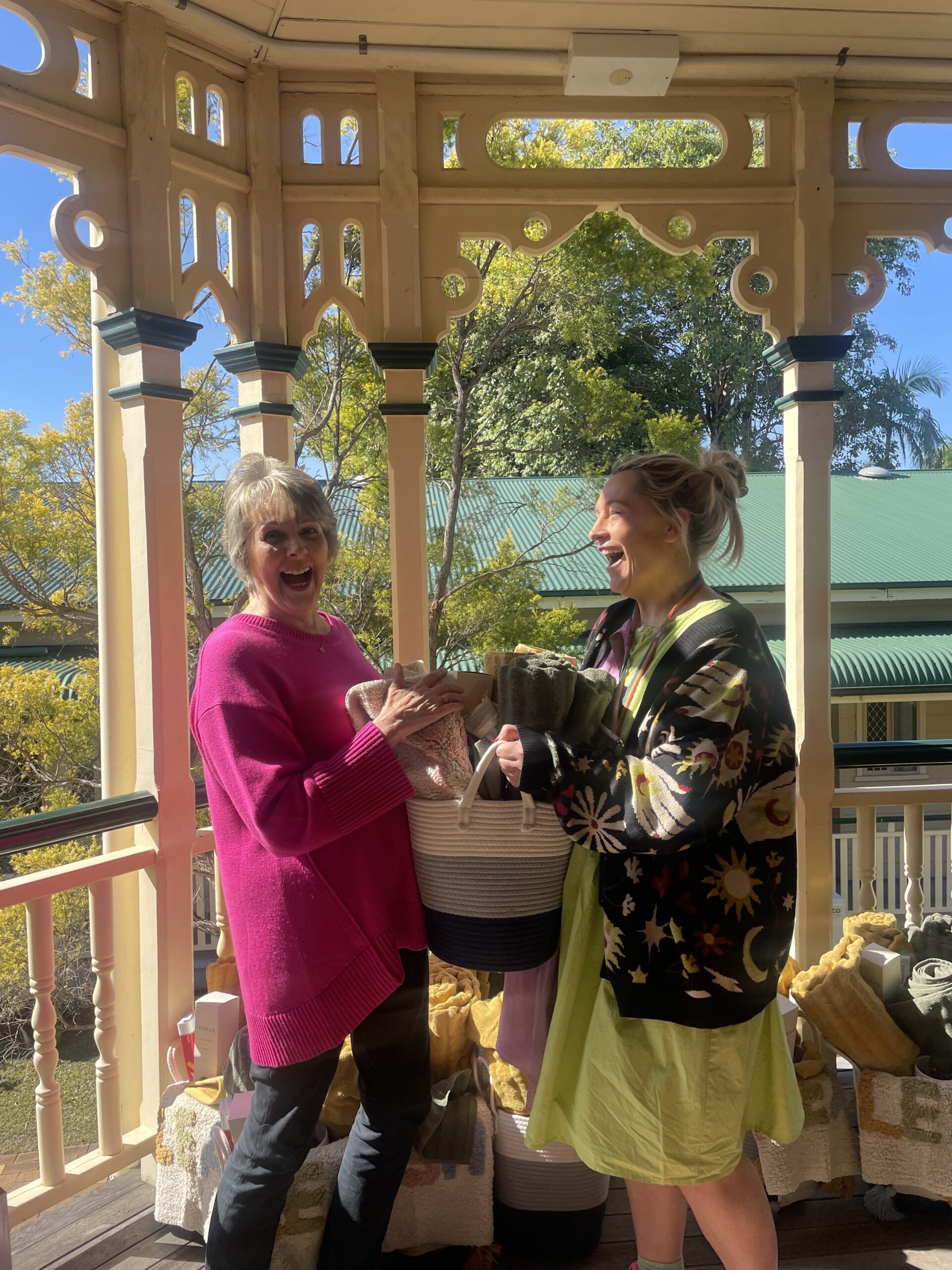 Two women joyfully holding a potted plant on a gazebo. One woman, with light skin, wears a bright pink sweater and has a cheerful expression. The other woman, with light skin, wears a black and yellow patterned cardigan and a light green dress, also smiling.