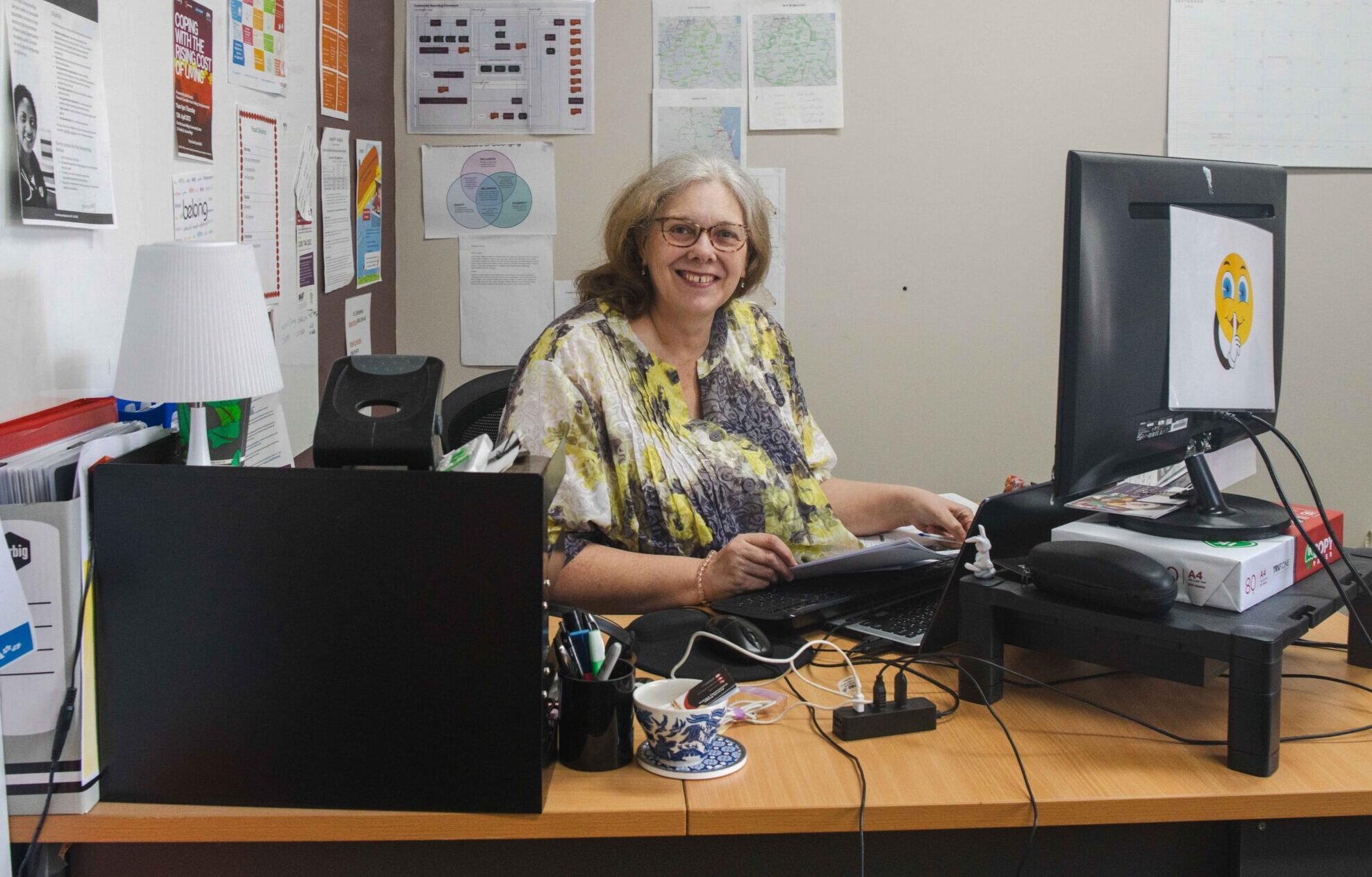 Woman sitting at a desk in an office, wearing a colorful floral blouse. She has light brown hair and is smiling while working on a computer. The workspace includes a black monitor and various documents on the wall.