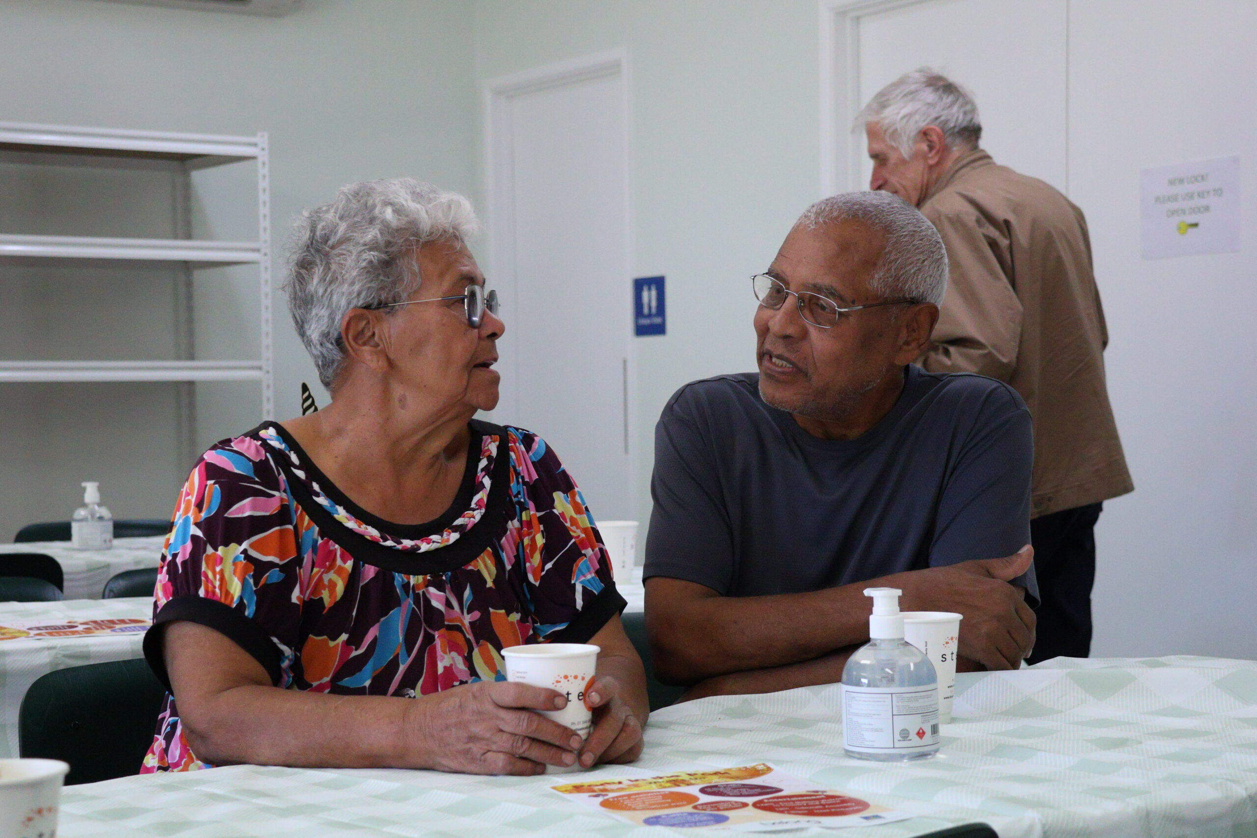 Two elderly individuals are engaged in conversation at a table. The woman, of Hispanic descent, wears a colorful floral blouse and has a friendly expression. The man, of African descent, is dressed in a dark gray shirt and appears to be smiling, showing interest in the discussion. In the background, an older man is seen standing.