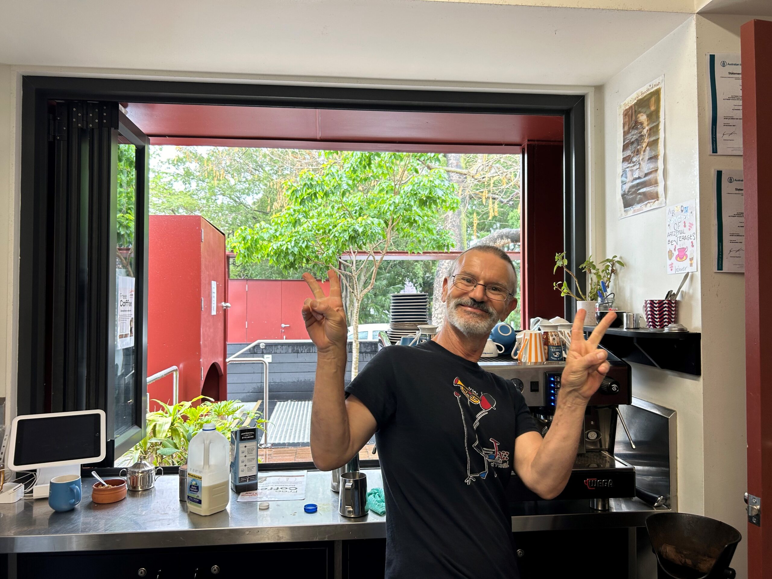 Smiling middle-aged man with a beard and glasses, wearing a black t-shirt, makes a peace sign in a café setting. Kitchen equipment and plants are visible in the background, along with a window showcasing greenery outside.