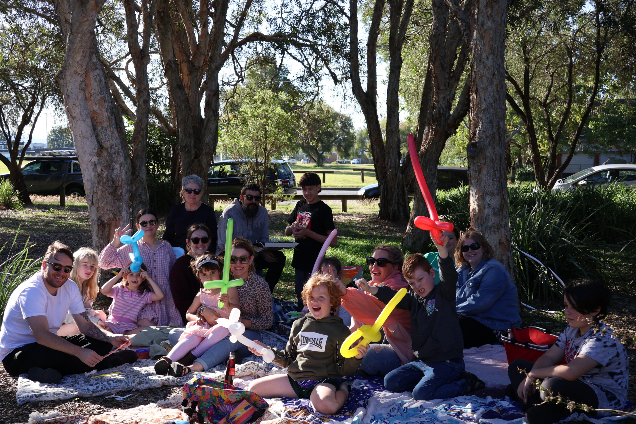 Group of people enjoying a picnic outdoors, featuring a diverse gathering. Some individuals are wearing casual clothing, including a black t-shirt and colorful balloon creations. The group consists of children and adults, with a mix of ethnicities. Several people are smiling and engaged, showcasing a festive atmosphere.