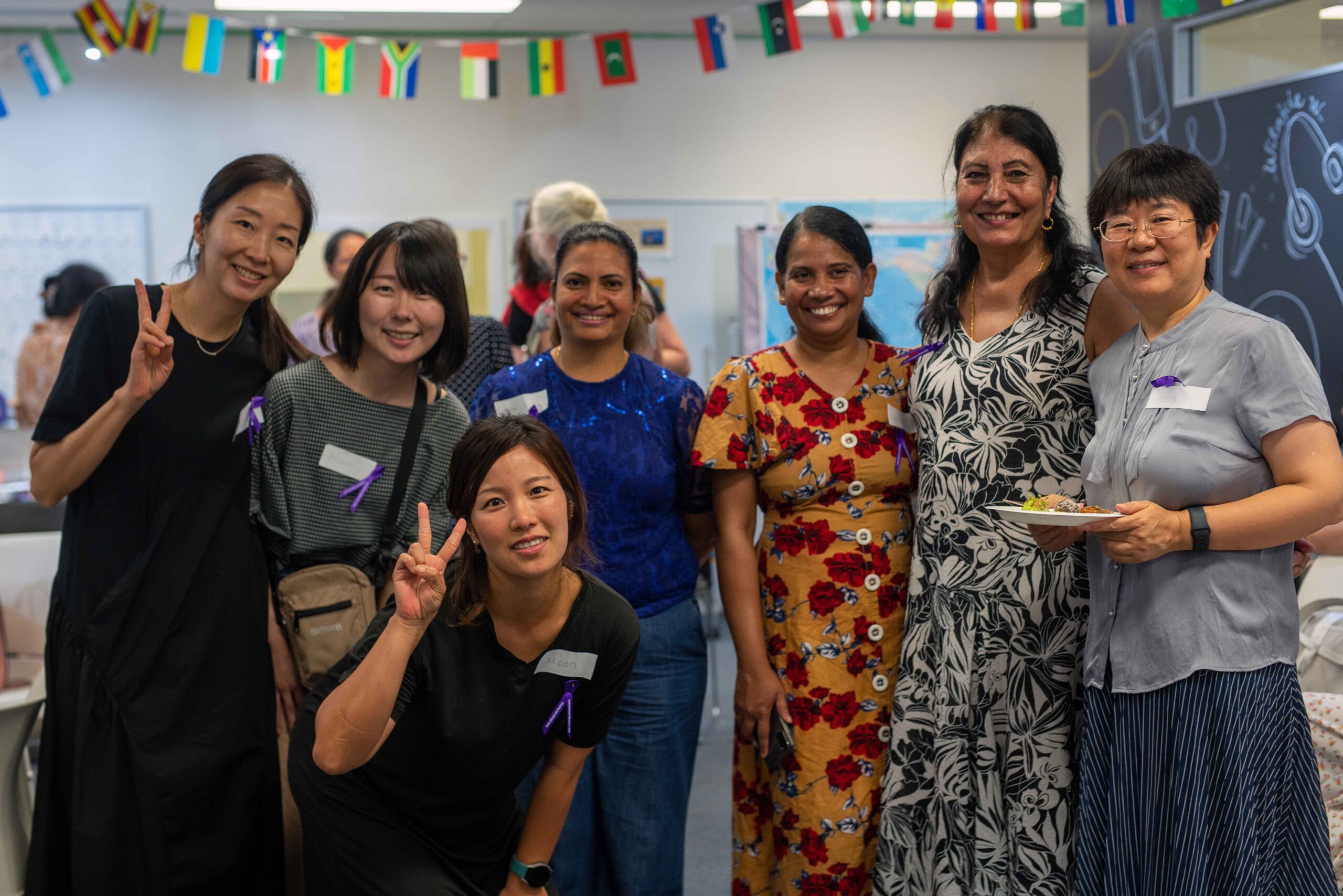 Group of diverse women smiling and posing together at a gathering. One woman in a black top and gray pants, another in a floral dress, and another in a blue top. They display a range of ethnicities and expressions of joy, with one woman making a peace sign.