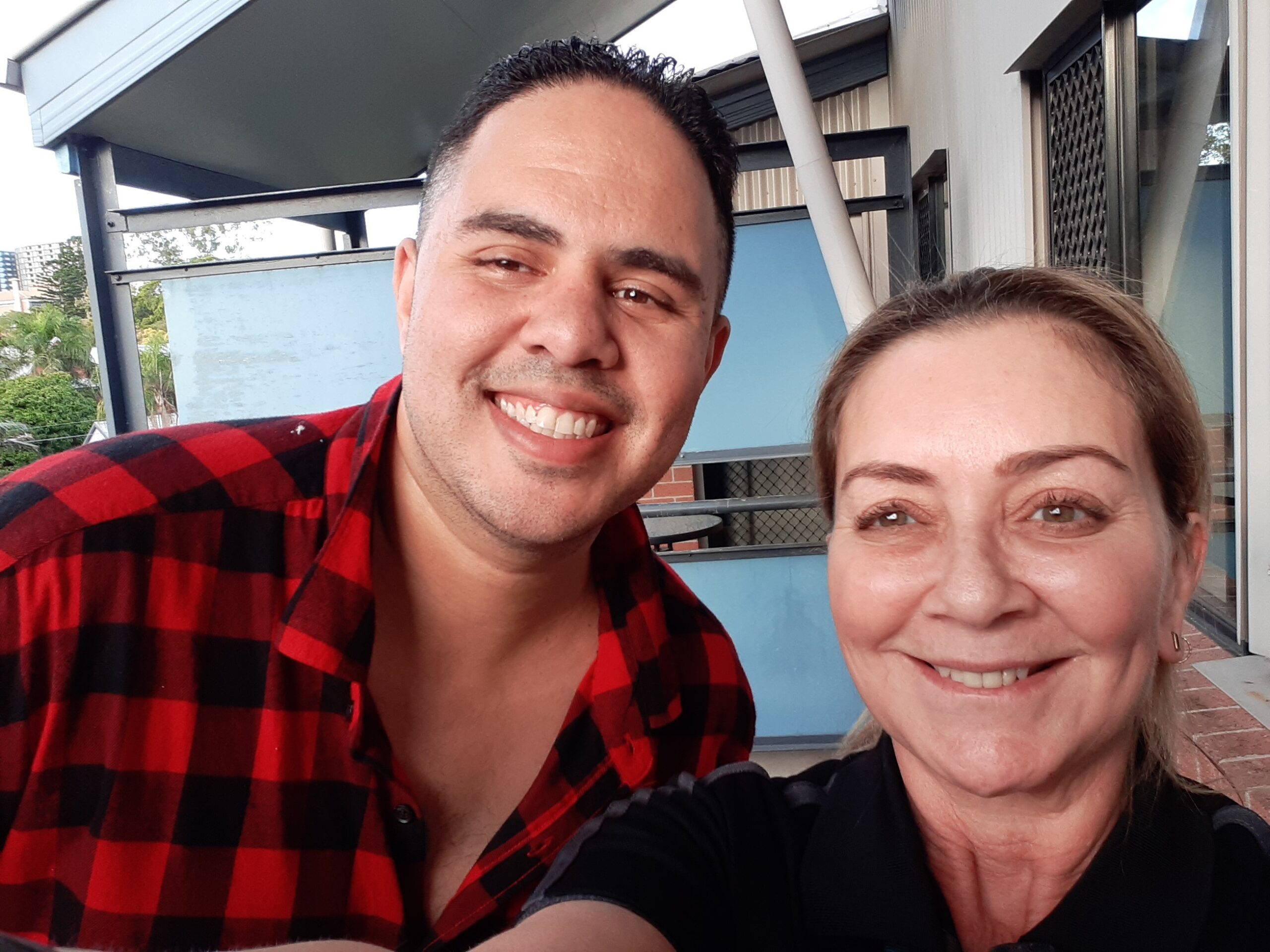 A man with a light brown complexion, wearing a red and black plaid shirt, smiles broadly alongside a woman with a fair complexion, wearing a black polo shirt. Both are taking a selfie, showcasing cheerful expressions.