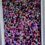 Varinia's artwork -Colorful abstract artwork featuring a multitude of vibrant beads and dots in shades of purple, pink, blue, green, and yellow, framed in an ornate silver frame. The composition showcases a dense arrangement of various shapes and sizes, creating a visually striking pattern.