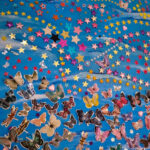 Colorful collage of butterflies and stars on a blue background, featuring various butterfly designs in shades of pink, yellow, and black, along with shiny star stickers in multiple colors, including gold, pink, and blue. The arrangement creates a whimsical and vibrant pattern.