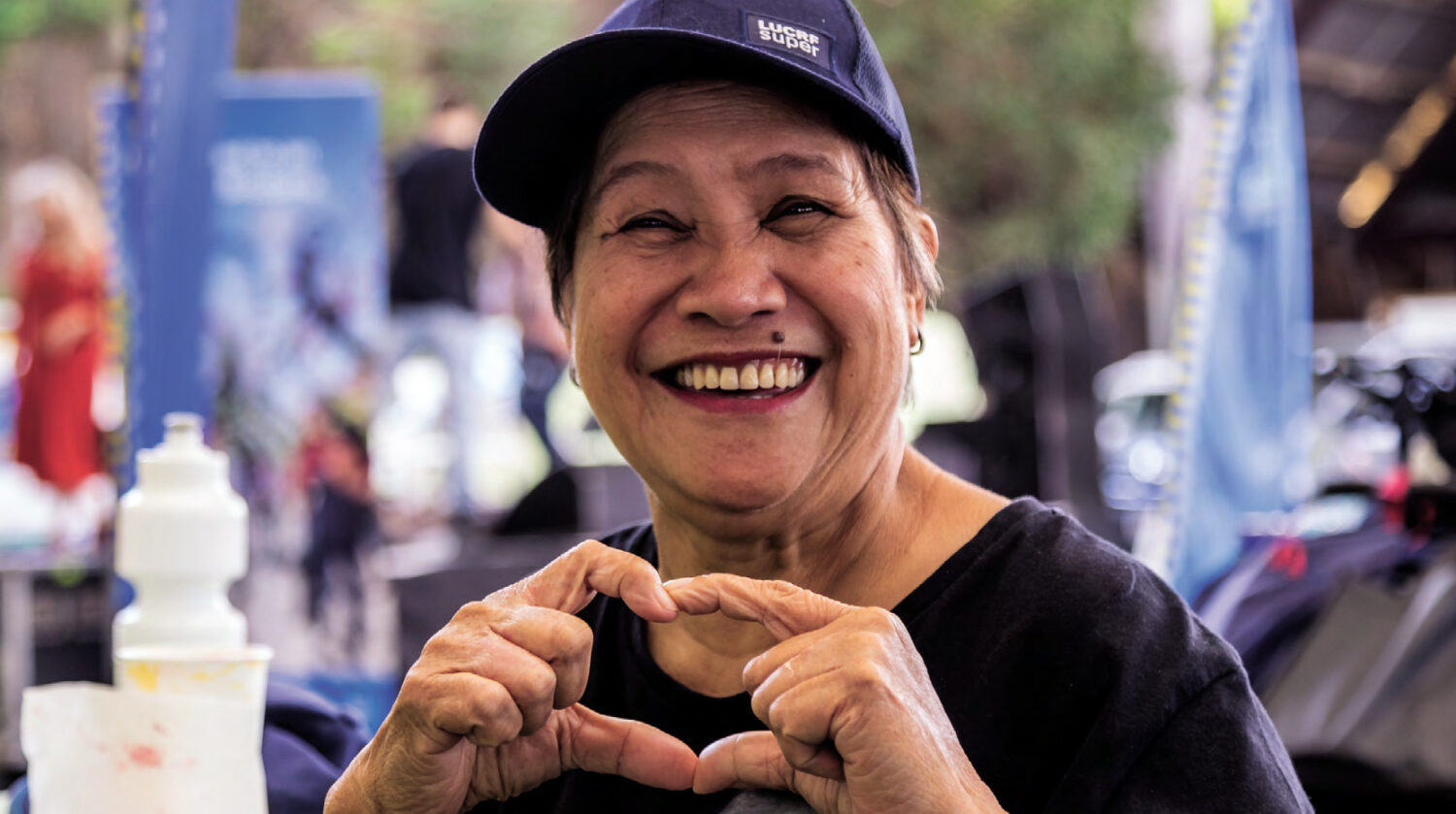 Smiling elderly woman making a heart shape with her hands, wearing a navy blue cap and a black shirt. The background features a blurred outdoor event setting, conveying a joyful and warm atmosphere.