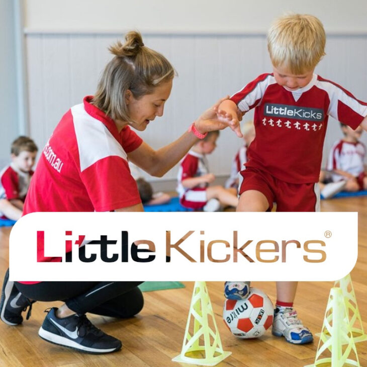 A female coach with light brown hair, wearing a red and white sports uniform, kneels beside a young boy with blonde hair, dressed in a red sports outfit. The boy is focused and smiling as he prepares to kick a soccer ball, with training cones in the background. The image promotes the Little Kickers program.