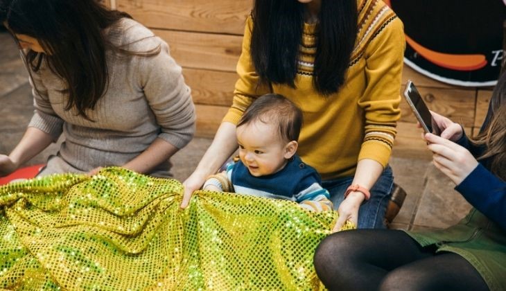 A joyful baby with light brown skin and short black hair plays with a shiny green sequin fabric, while two adults assist him. One adult, wearing a yellow sweater, has long black hair and an Asian ethnicity, and the other, in a beige sweater, has medium-length brown hair and appears to be of Caucasian descent.