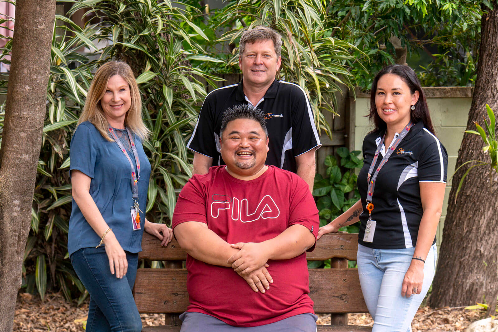 Group of four individuals posing outdoors. A Caucasian woman on the left wearing a blue shirt, a Caucasian man in the back center wearing a black shirt, an Asian man in the front wearing a red FILA shirt, and a Hispanic woman on the right wearing a black and white shirt. All are smiling and appearing friendly.