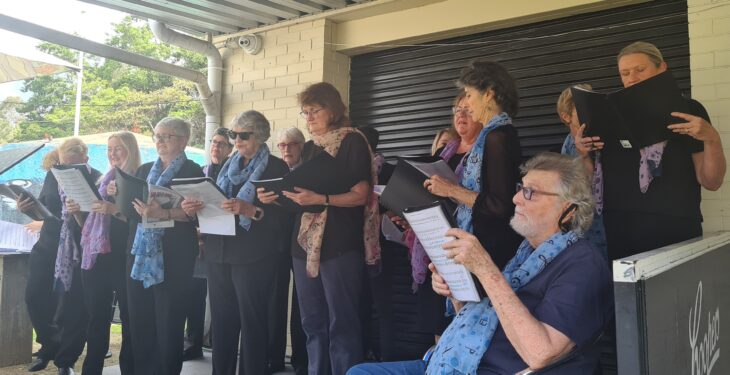 A group of diverse individuals singing together outdoors. Most are wearing black clothing, with some featuring blue scarves. They display various expressions of focus and enjoyment while holding sheet music.