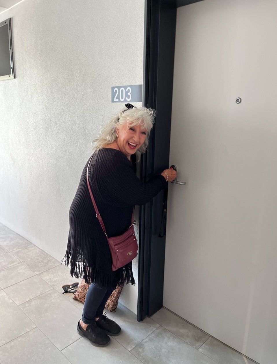 A woman with light skin and blonde hair is smiling broadly while unlocking a door; She is wearing a black sweater and a red crossbody bag, with a playful expression on her face.