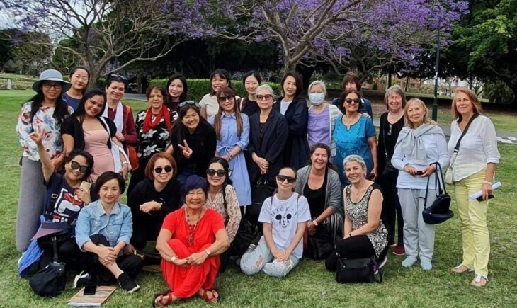 Group of diverse women posing together outdoors, wearing a variety of outfits including red, white, and blue clothing. Some women are smiling while others are serious. The group includes individuals of different ethnicities, showcasing a range of hairstyles and accessories, such as sunglasses and hats.