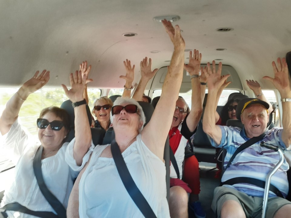 Group of diverse individuals in a vehicle, all wearing seatbelts and raising their hands enthusiastically. Women in the front, one wearing a white top and sunglasses, while others have various outfits including a red shirt and a striped top. Their expressions show joy and excitement.