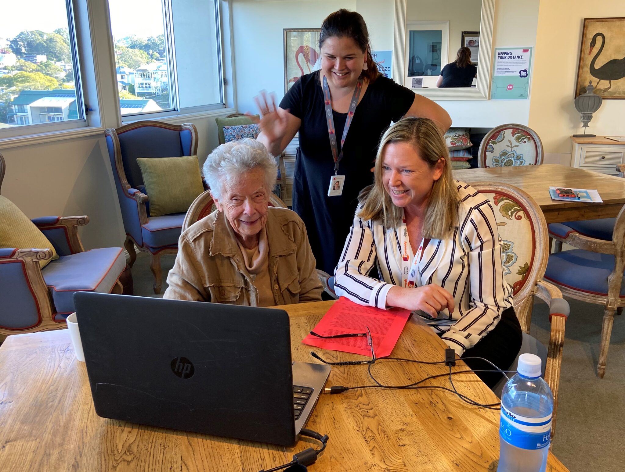 Three women are gathered around a laptop at a wooden table. An elderly woman with gray hair, wearing a brown cardigan, appears engaged as she looks at the screen. A middle-aged woman with blonde hair, dressed in a striped blouse, smiles while pointing at the laptop. Behind them, a younger woman with dark hair, wearing a black shirt, is playfully waving her hand.