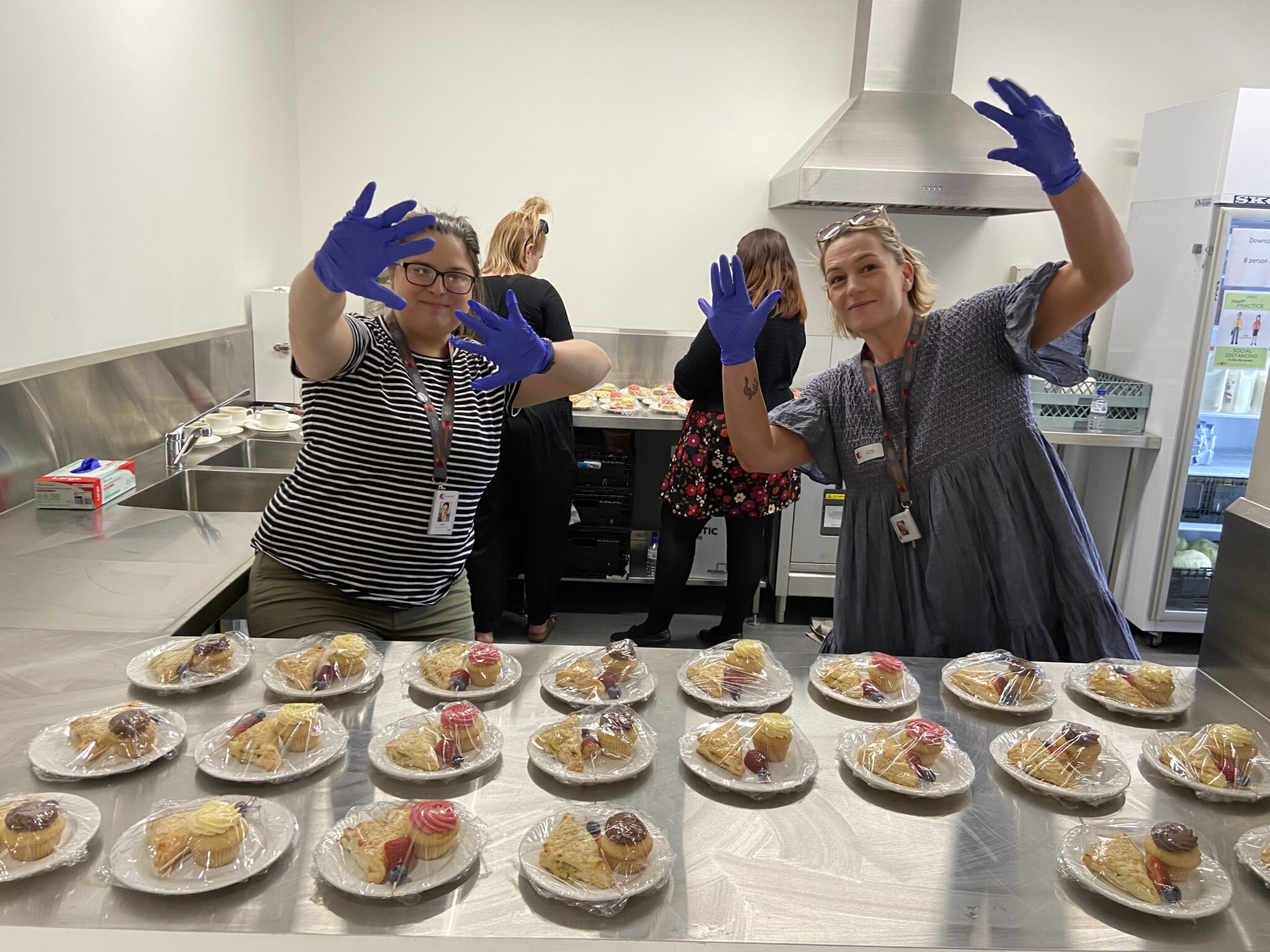 Two women in a kitchen wearing blue gloves and smiling enthusiastically, with one in a gray striped shirt and the other in a gray shirt. They are posing playfully in front of a table filled with plates of food. In the background, another individual is visible, wearing a black outfit with a floral pattern.