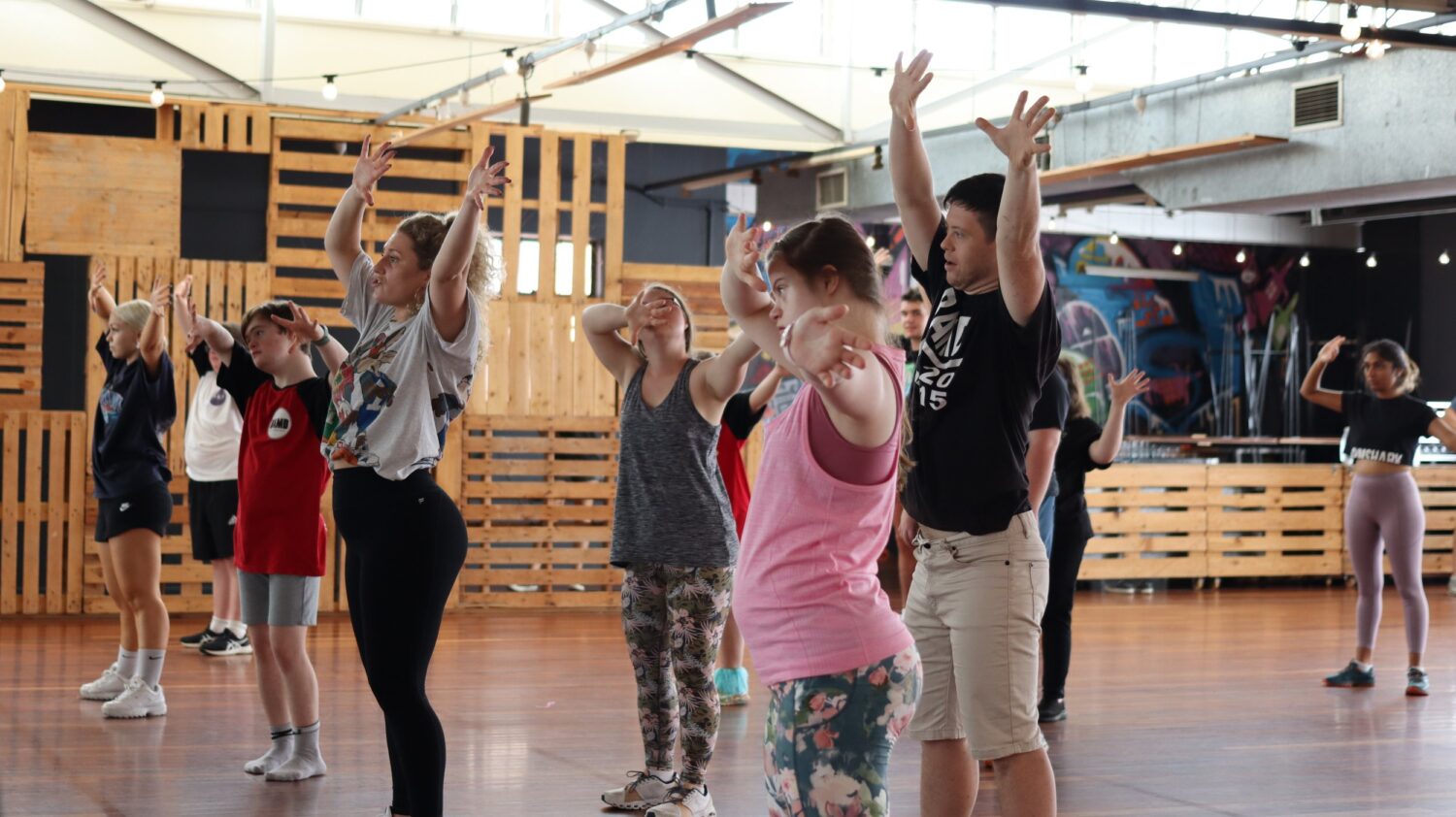 Group of diverse dancers in a studio, performing a routine. The instructor is in a pink tank top and patterned leggings, looking focused. Others wear a mix of casual clothing, including t-shirts and shorts, with various hair colors and styles. The group is engaged and expressive, with arms raised.