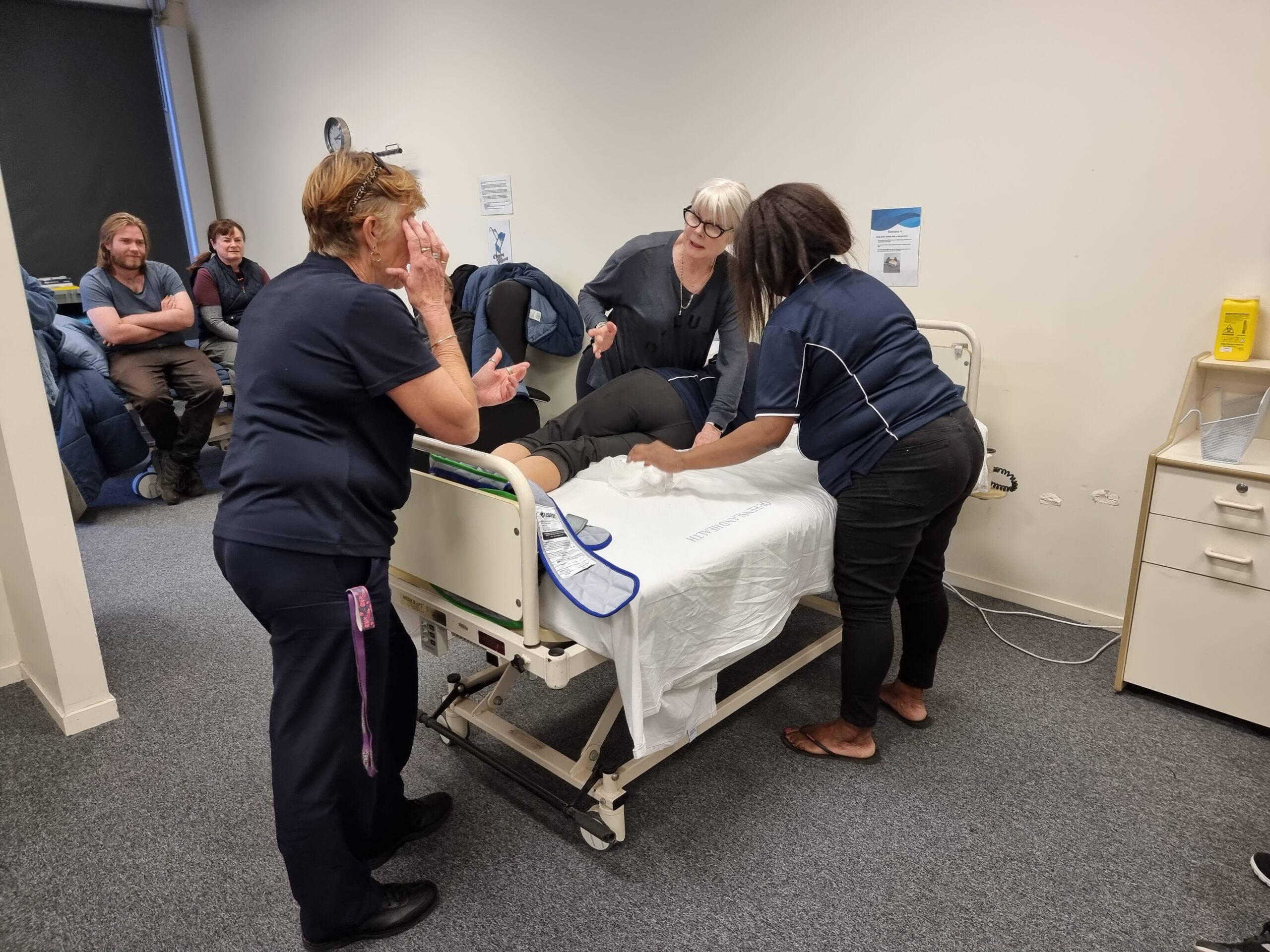 Two healthcare professionals, are demonstrating how to turn a patient over to a group of students.