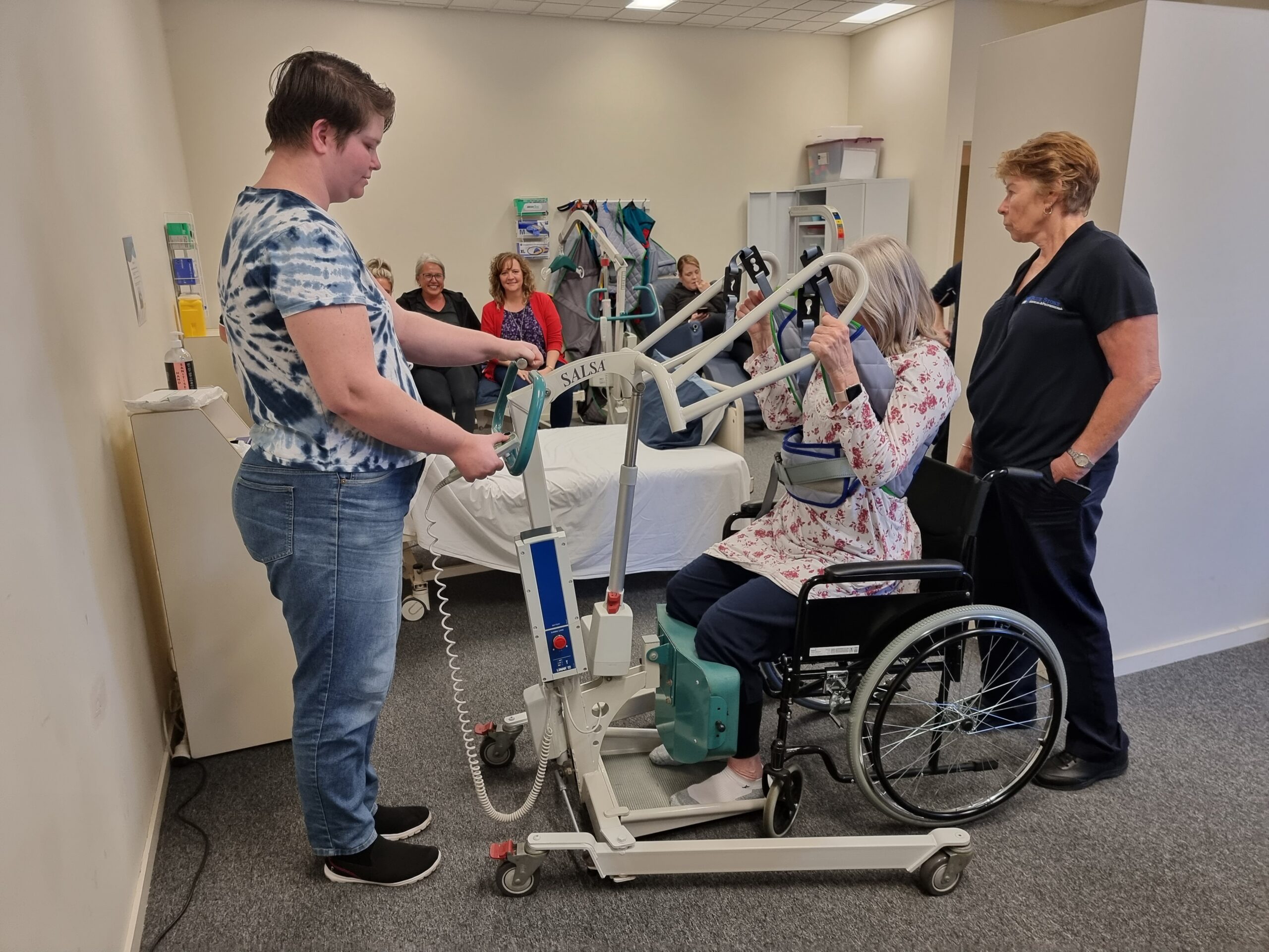 A person with short dark hair, wearing a patterned blue shirt and jeans, operates a patient lift. In the wheelchair, a woman with light skin and gray hair, appears focused as she prepares for transfer. A woman with light skin and brown hair, wearing a black shirt, observes the process. A group of students is sitting at the back of the room observing the demonstration