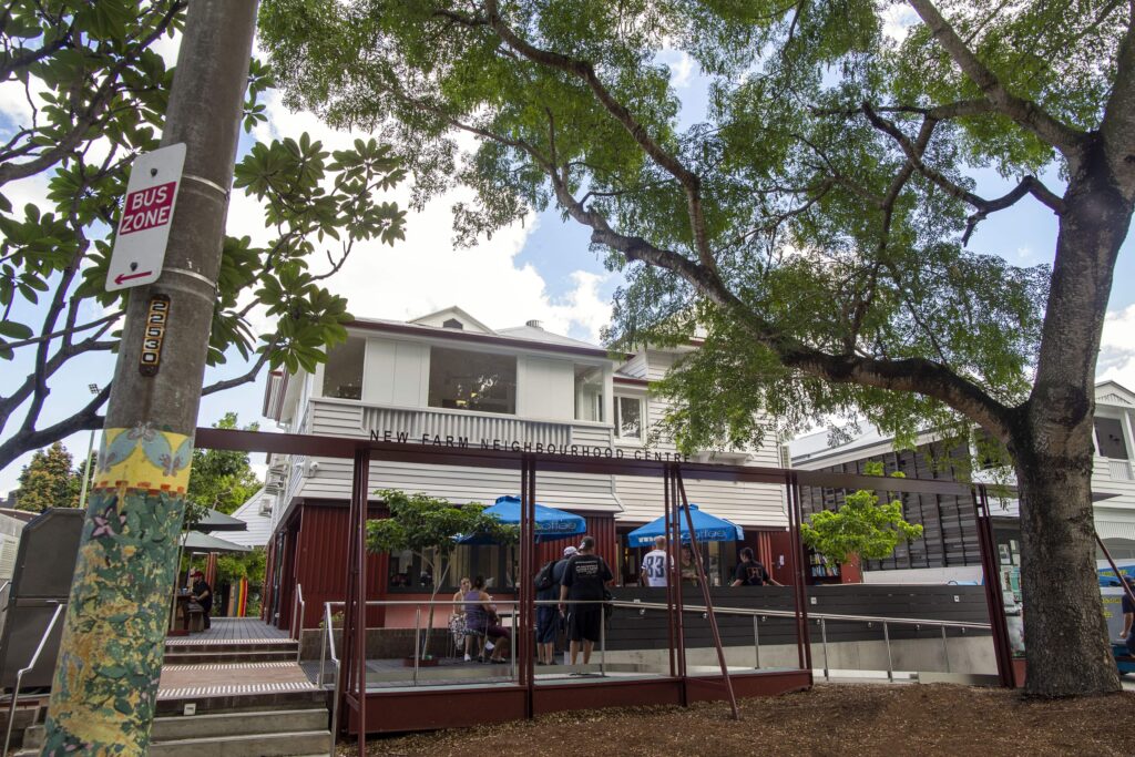 Image of a two-story building with a large tree in the foreground, featuring outdoor seating areas with blue umbrellas. There are several people, including a person wearing a black shirt and another in casual attire, engaged in conversation. The setting appears to be a communal area.