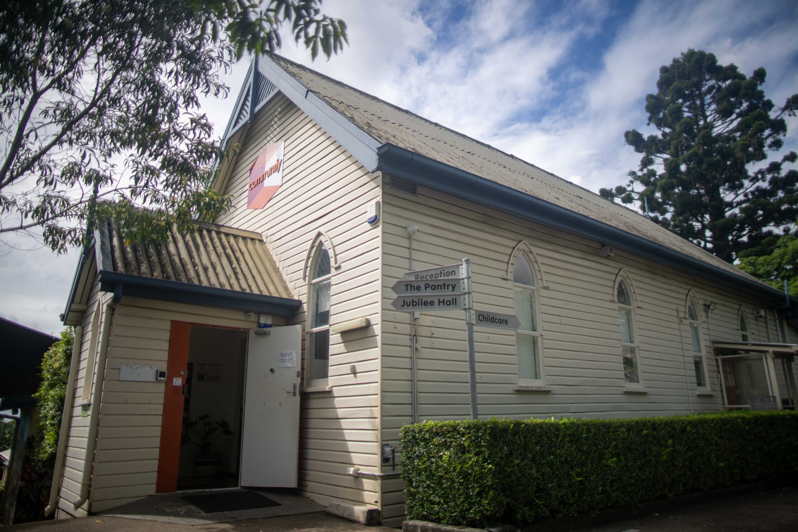 Image of a white wooden church building featuring a peaked roof and arched windows. A sign near the entrance indicates the church name. Green shrubs and trees are visible in the foreground.