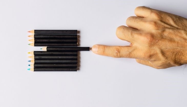 A hand with a light skin tone is gently touching a black pencil while resting on a row of black pencils arranged on a white surface.