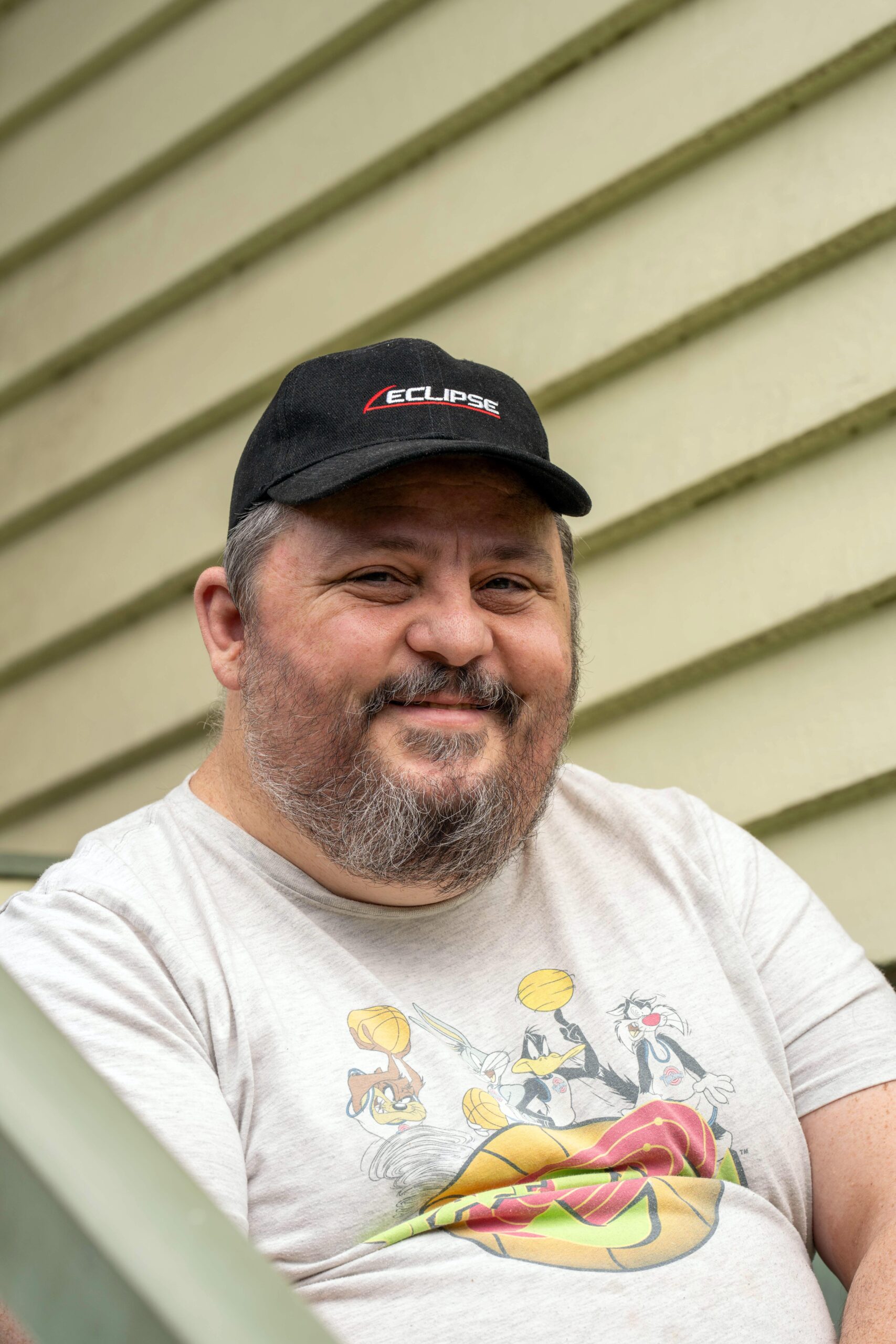 Photo of Peter - Smiling middle-aged man with a beard, wearing a black cap and a light-colored t-shirt featuring cartoon characters, sitting against a green wall.