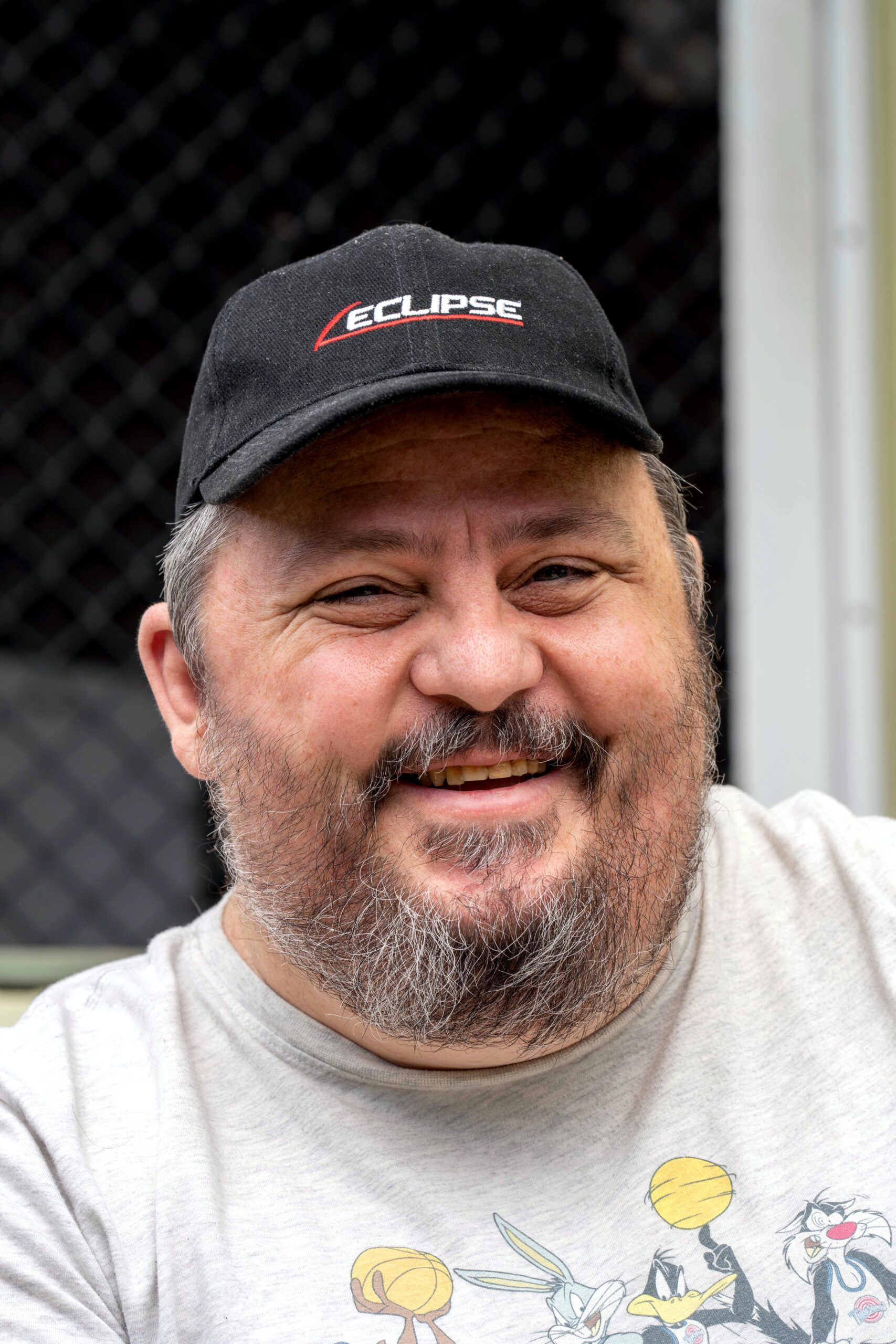 Smiling man with a beard wearing a black cap and a light gray t-shirt, with a cheerful expression as he looks at the camera