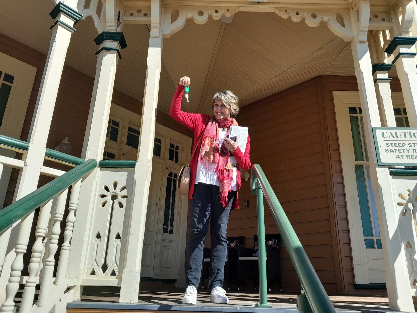 An older woman is smiling, and standing at the top of a staircase, near the front door of a house, holding up a set of keys