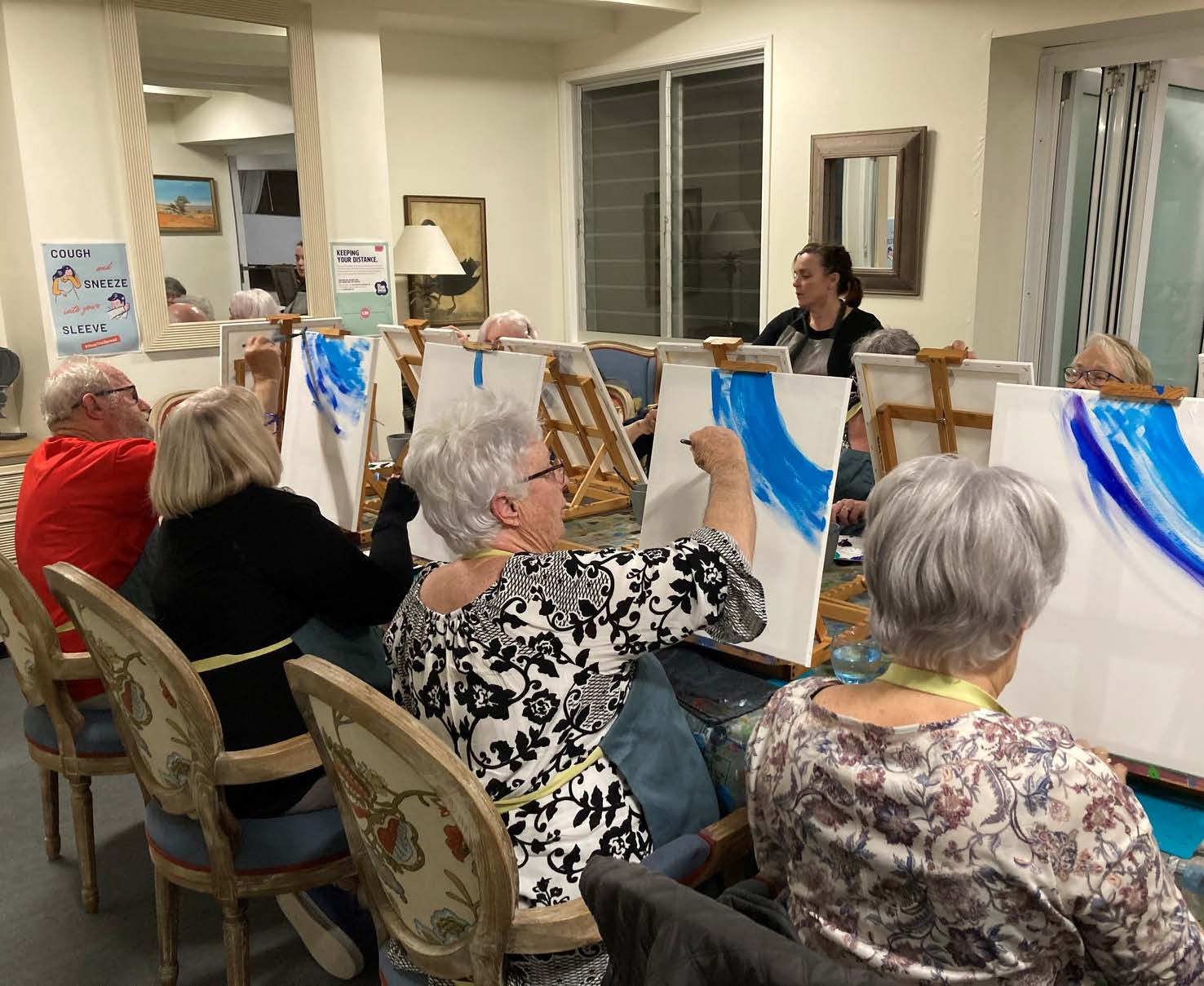 A group of older women engaged in a painting class, seated at easels. One woman with dark hair and a black top instructs the class, while participants wear various outfits including a red sweater, a patterned black and white shirt, and several with gray hair. The women appear focused as they paint blue designs on their canvases.