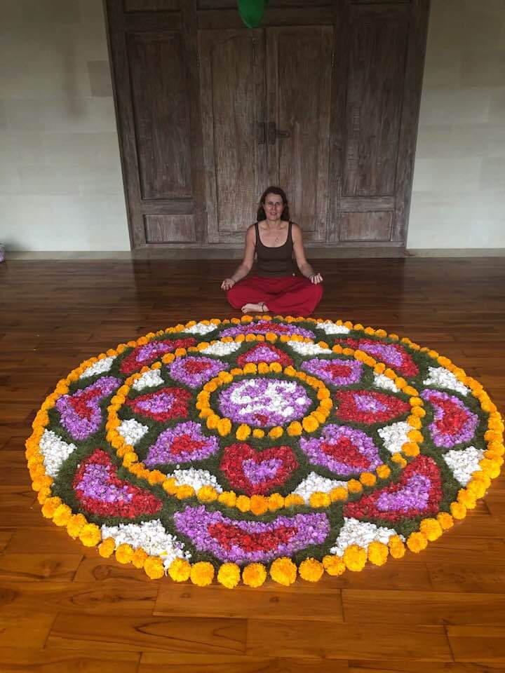 A woman with long dark hair sits cross-legged on a wooden floor, wearing a black tank top and red pants, in front of a vibrant flower mandala. The mandala features an array of colorful flowers, including pink, white, orange, and yellow, arranged in a circular pattern.