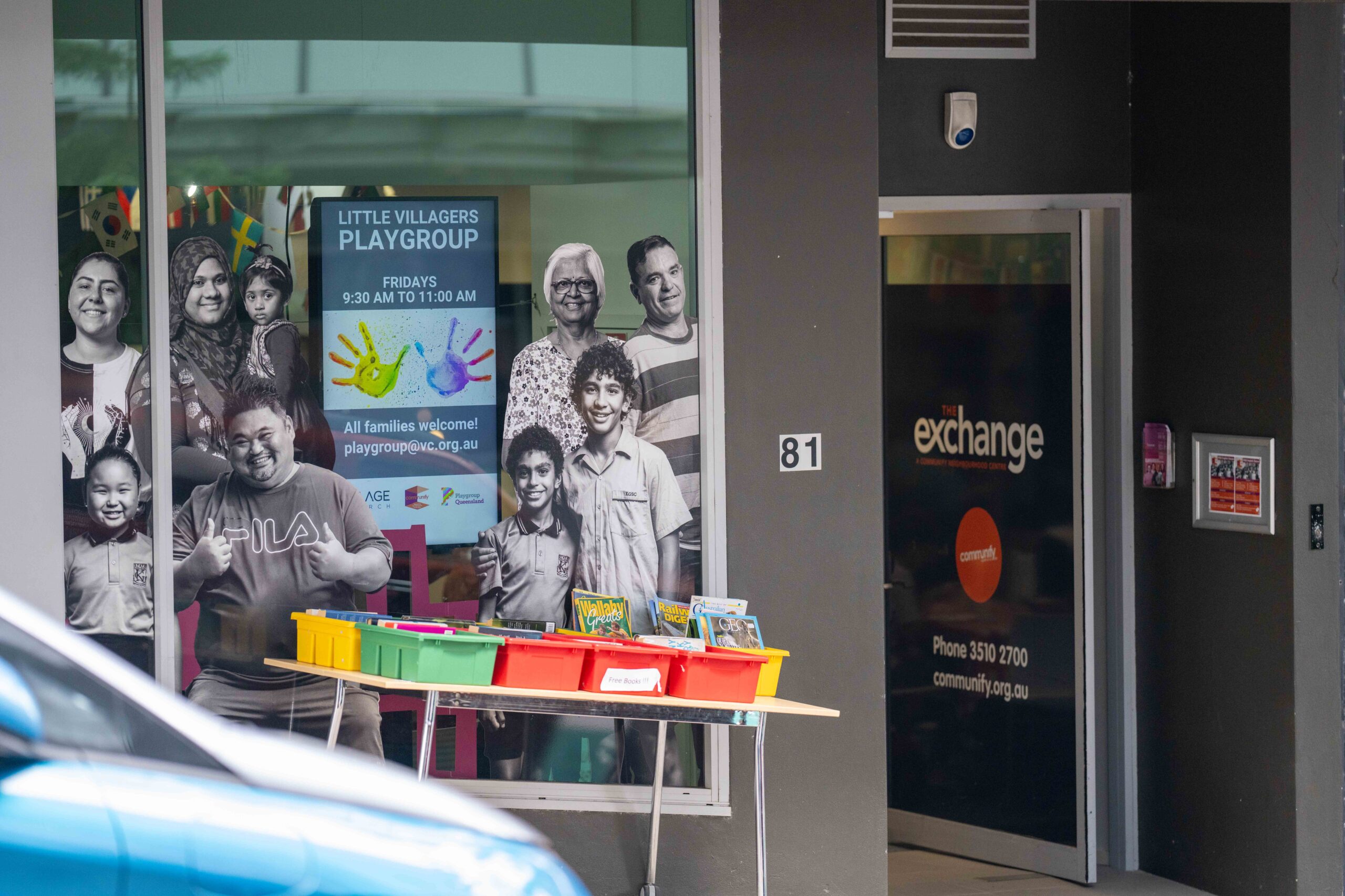 A colorful play area with a table displaying various toys and art supplies is visible through the window of a building. In the background, black-and-white photographs of diverse individuals, including adults and children, are featured on the wall. The entrance door displays the word "exchange" prominently.