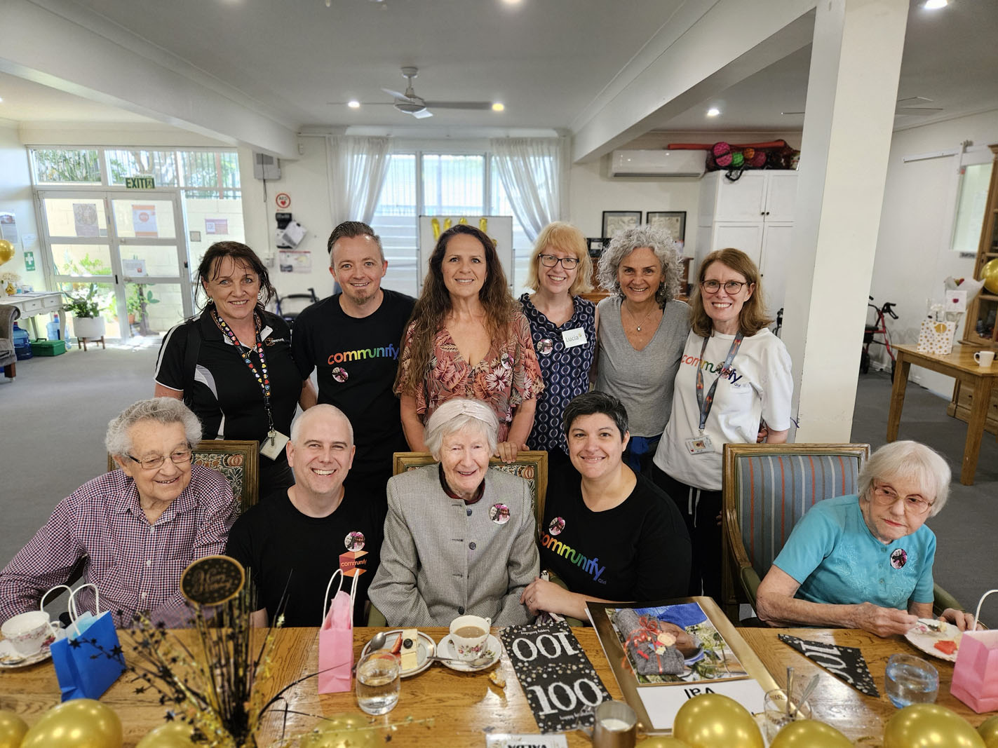 Group photo of a diverse team and residents at a community event. The front row features older women in various outfits, including a patterned purple top and a light grey jacket. In the back row, individuals wear black shirts, white shirts, and colorful attire, displaying smiles. The group is gathered around a table decorated with balloons and magazines.