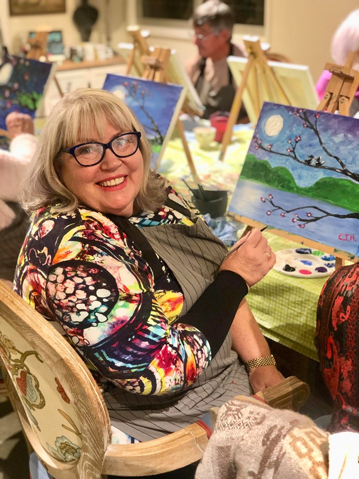 Smiling woman with gray hair and glasses, wearing a colorful patterned cardigan, holding a paintbrush in front of her canvas. The background features other artists working on their paintings.