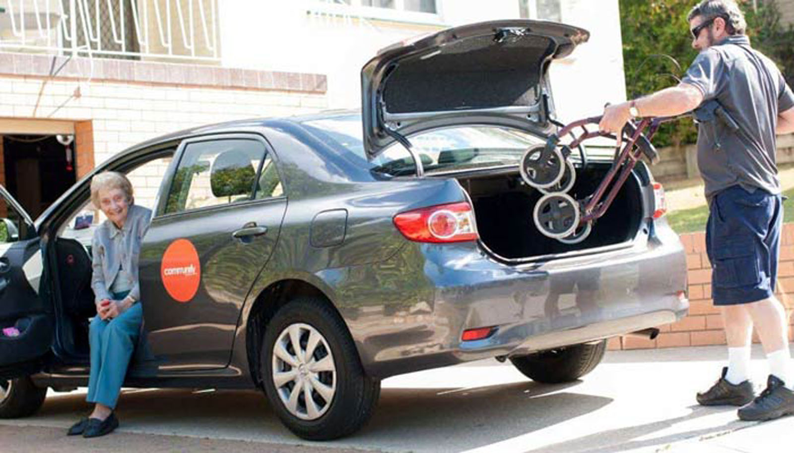 A person loading a mobility walker into the trunk of a dark blue sedan. There is an elderly lady waiting in the passenger seat, smiling at the camera