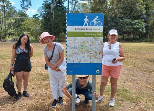 Group of four individuals at a park sign, indicating walking and biking paths. Two women of Asian descent, one wearing a black outfit and the other in white pants and a pink hat, smile at the camera. A man of Asian descent squats down wearing a blue shirt and jeans. Another woman, of Caucasian descent, stands beside them in a white shirt and pink shorts, also smiling.