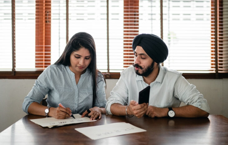 A South Asian woman with long dark hair, wearing a light blue shirt, is focused on writing on a piece of paper. A South Asian man with a beard, wearing a black turban and a light gray shirt, is looking at her while holding a smartphone. They are seated at a wooden table with documents in front of them.