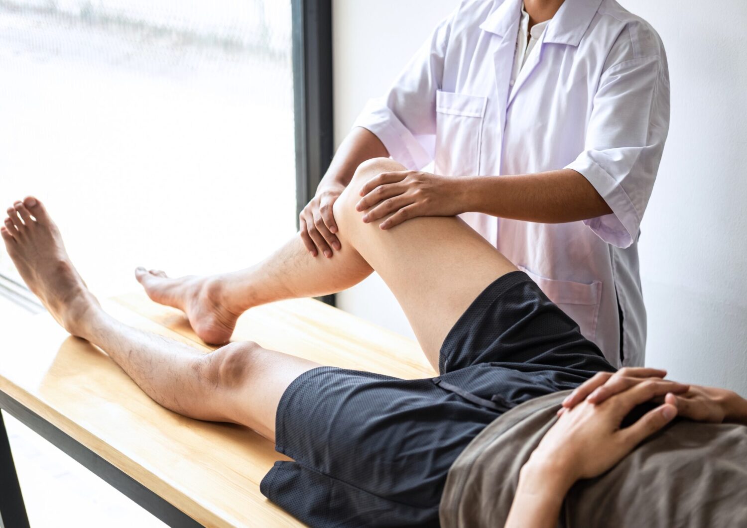 A healthcare professional in a white lab coat assists a patient with leg rehabilitation. The patient, wearing dark shorts and a gray shirt, appears relaxed as the therapist gently supports their leg. This image illustrates physical therapy and rehabilitation practices.