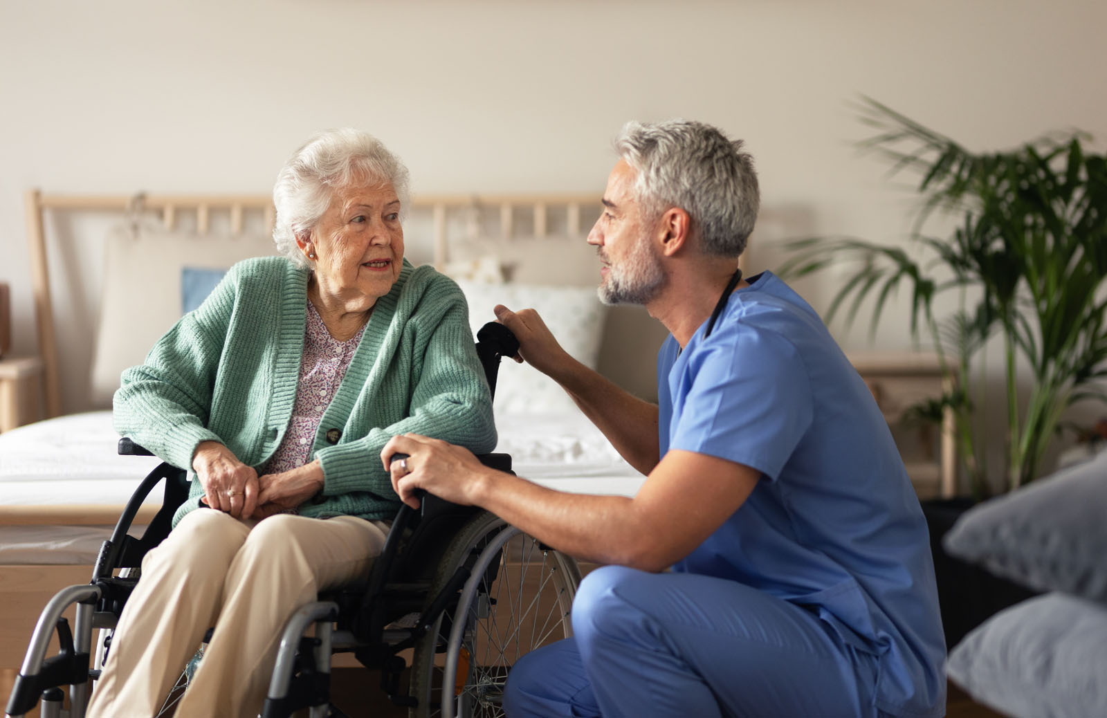 A healthcare professional in blue scrubs interacts with an elderly woman in a wheelchair. The woman, with gray hair and wearing a green cardigan, smiles gently at the caregiver, who has a focused expression.