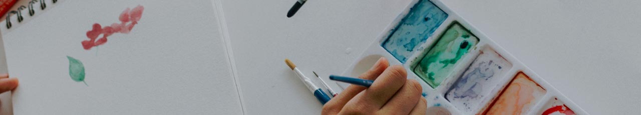 Close-up of a hand holding a blue paintbrush, with a yellow-tipped brush and a fine-tip brush nearby, on a white surface. The image focuses on art supplies, highlighting tools used for painting or drawing.