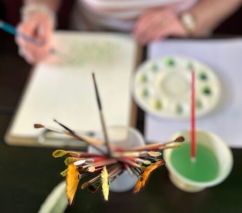 Art supplies including paintbrushes in various colours, and a palette with paint dots are arranged in focus, with a person's hands holding a pencil and a sketchbook in the background.