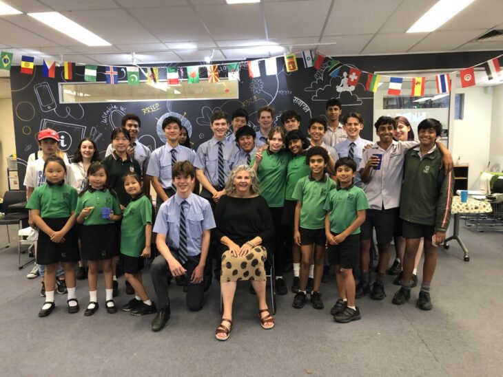 Group of students and a teacher in a classroom setting, with students wearing green uniforms and a teacher in a black top with a polka dot skirt. The group includes both boys and girls of diverse ethnicities, smiling and posing together.