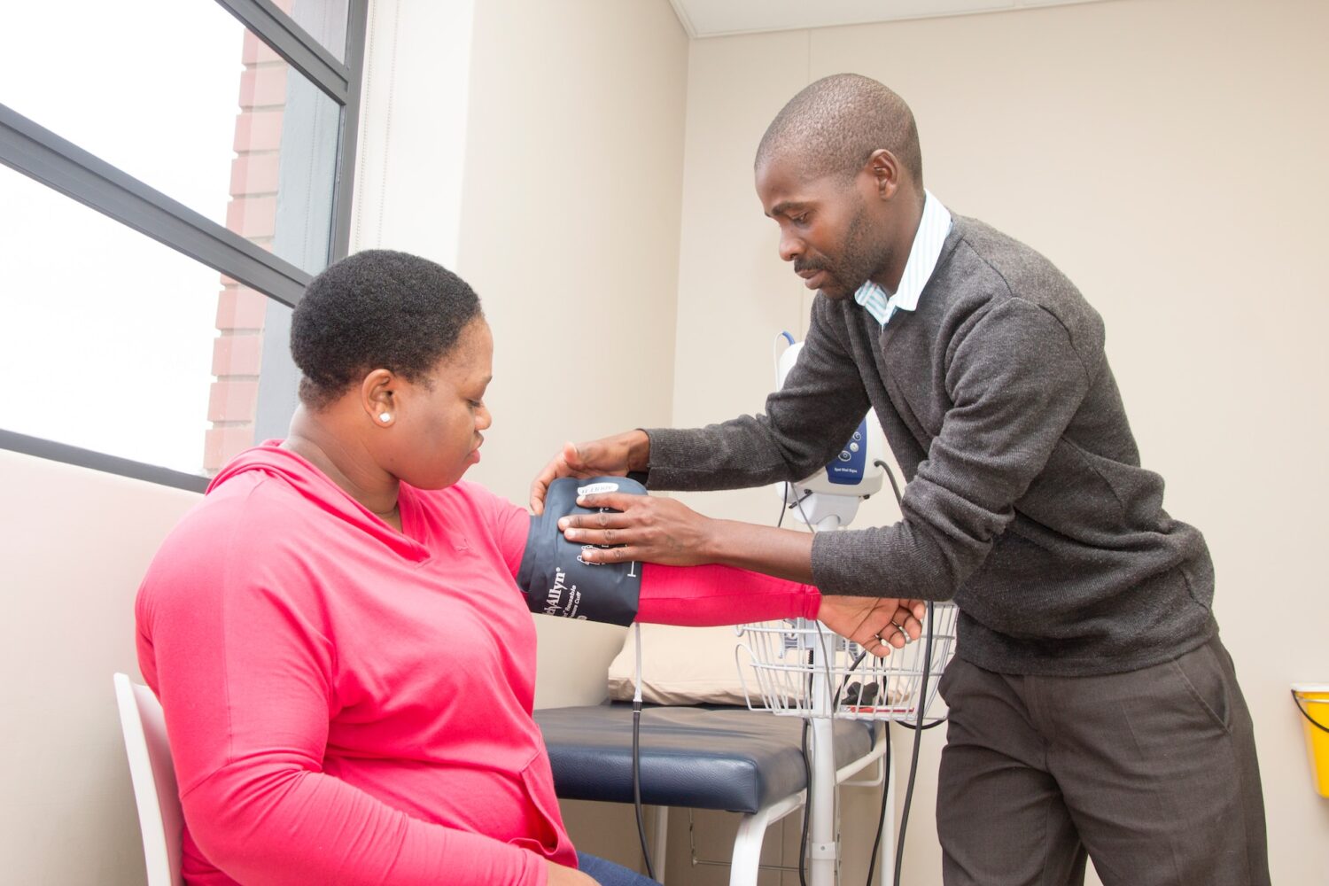 A healthcare professional, wearing a gray sweater and white shirt, is taking the blood pressure of a woman seated in a pink hoodie. The woman appears calm and attentive as the procedure is being conducted in a medical setting.