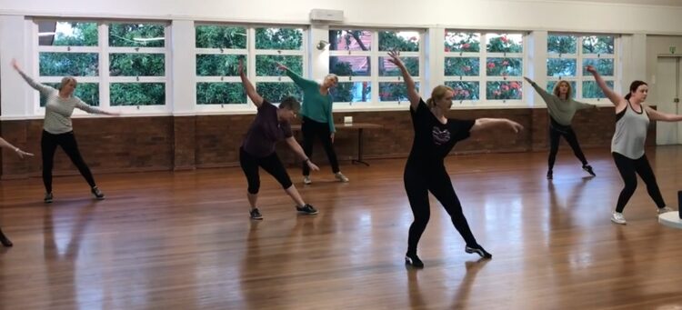 Three women are dancing in a spacious studio. The woman on the left, wearing a dark purple top and black leggings, appears focused. The woman in the center is dressed in a teal top and black pants, showing a joyful expression. The woman on the right is in a black shirt and leggings, demonstrating an enthusiastic pose.