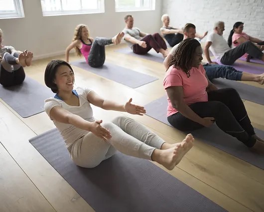 Group of diverse individuals practicing yoga in a bright studio. A woman of Asian descent in a white top and light pants is smiling while performing a seated pose, showcasing focus and relaxation. Other participants are engaged in various yoga positions on mats. The atmosphere is calm and supportive.